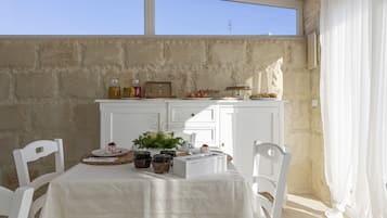 Un comedor con una mesa preparada para dos personas, un armario blanco con alimentos, y vista al cielo a través de una ventana.