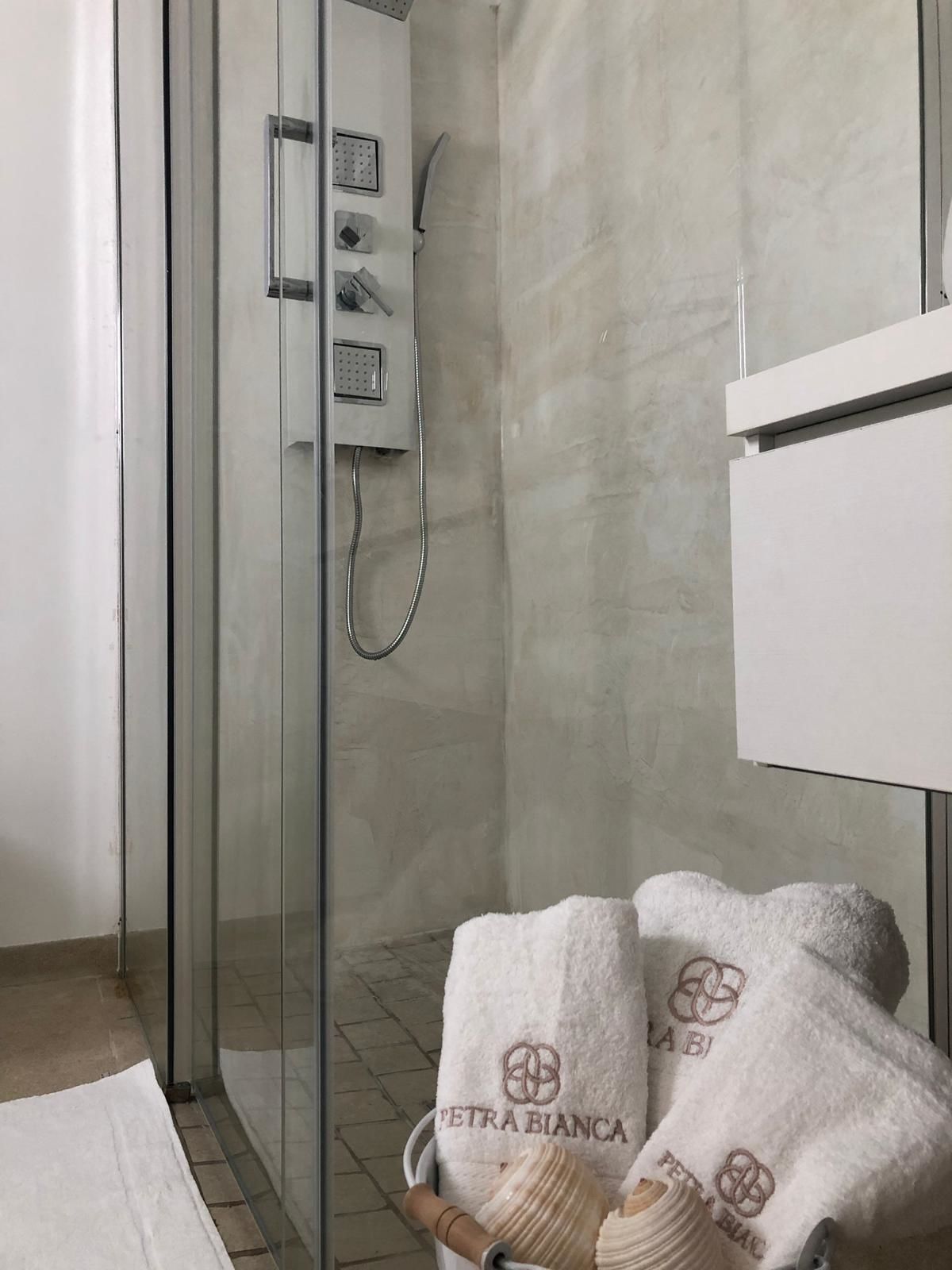 A modern bathroom with a glass shower enclosure, a white towel with 'PETRA BIANCA' branding, and a wooden bath mat.