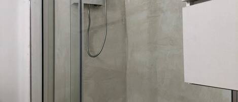 A modern bathroom with a glass shower enclosure, a white towel with 'PETRA BIANCA' branding, and a wooden bath mat.