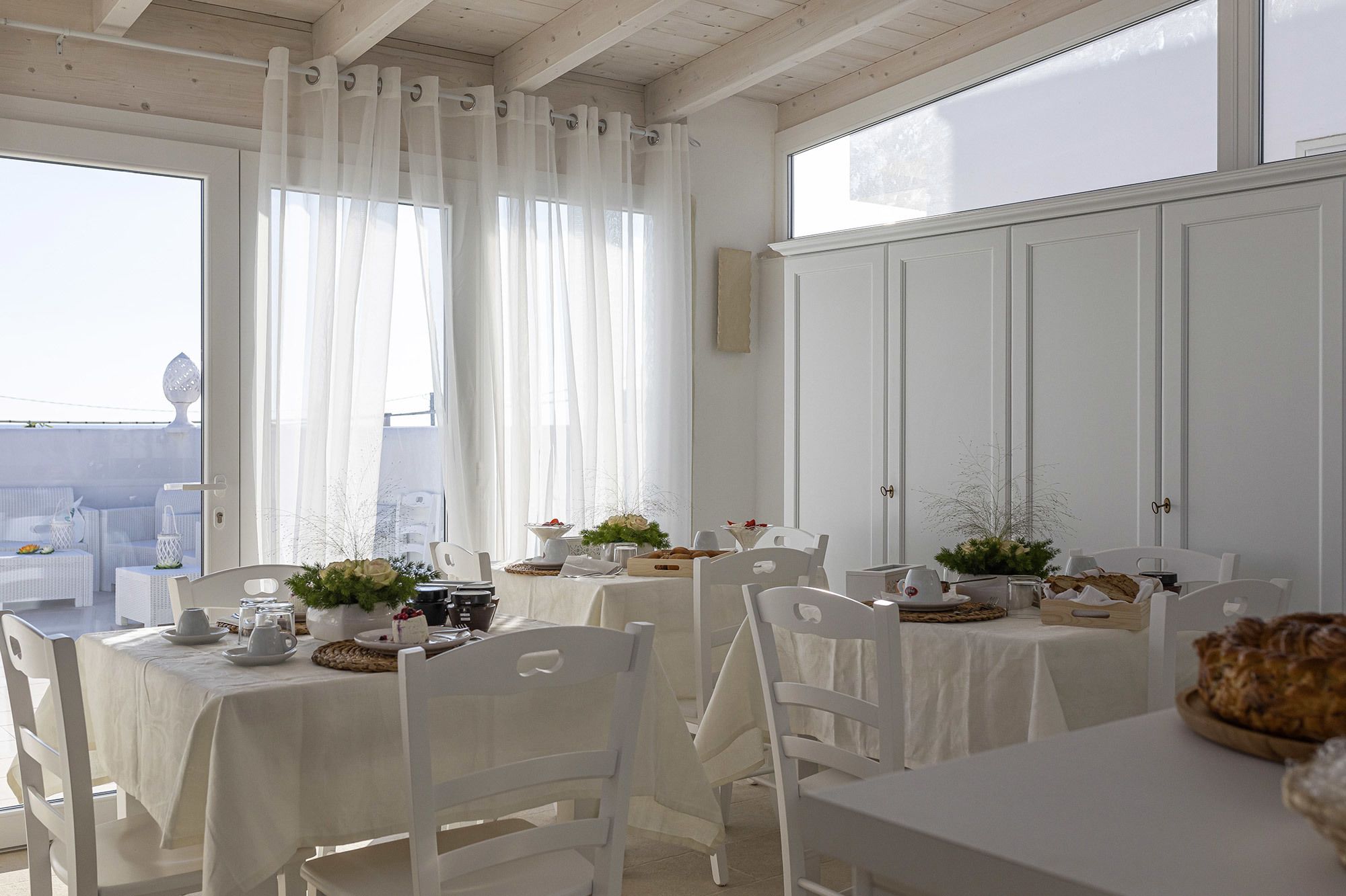 A dining area with white tablecloths, white chairs, and a view of the sea through large windows.
