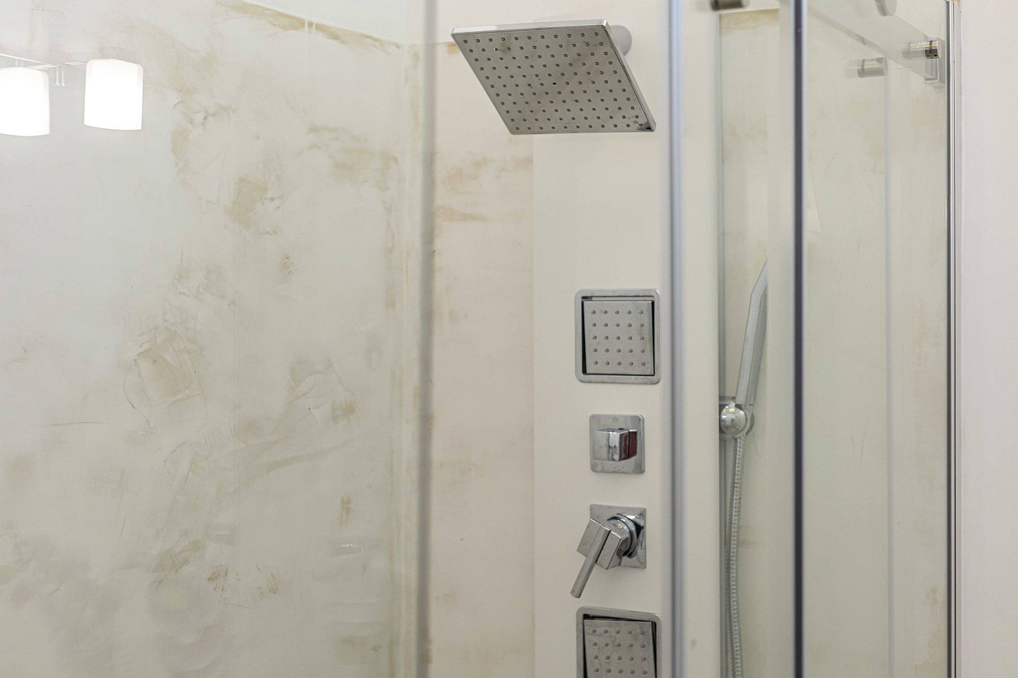 A glass-enclosed shower with a handheld showerhead, a wall-mounted control panel, and a square showerhead.
