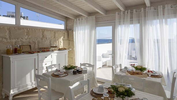 A dining area with white furniture, a buffet table with food and drinks, and a view of the sea through large windows. - Petra Bianca (Ugento)