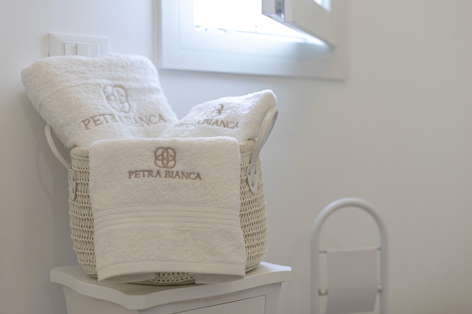 A white chair with a woven backrest and a towel draped over it, both branded with "PETRA BIANCA".
