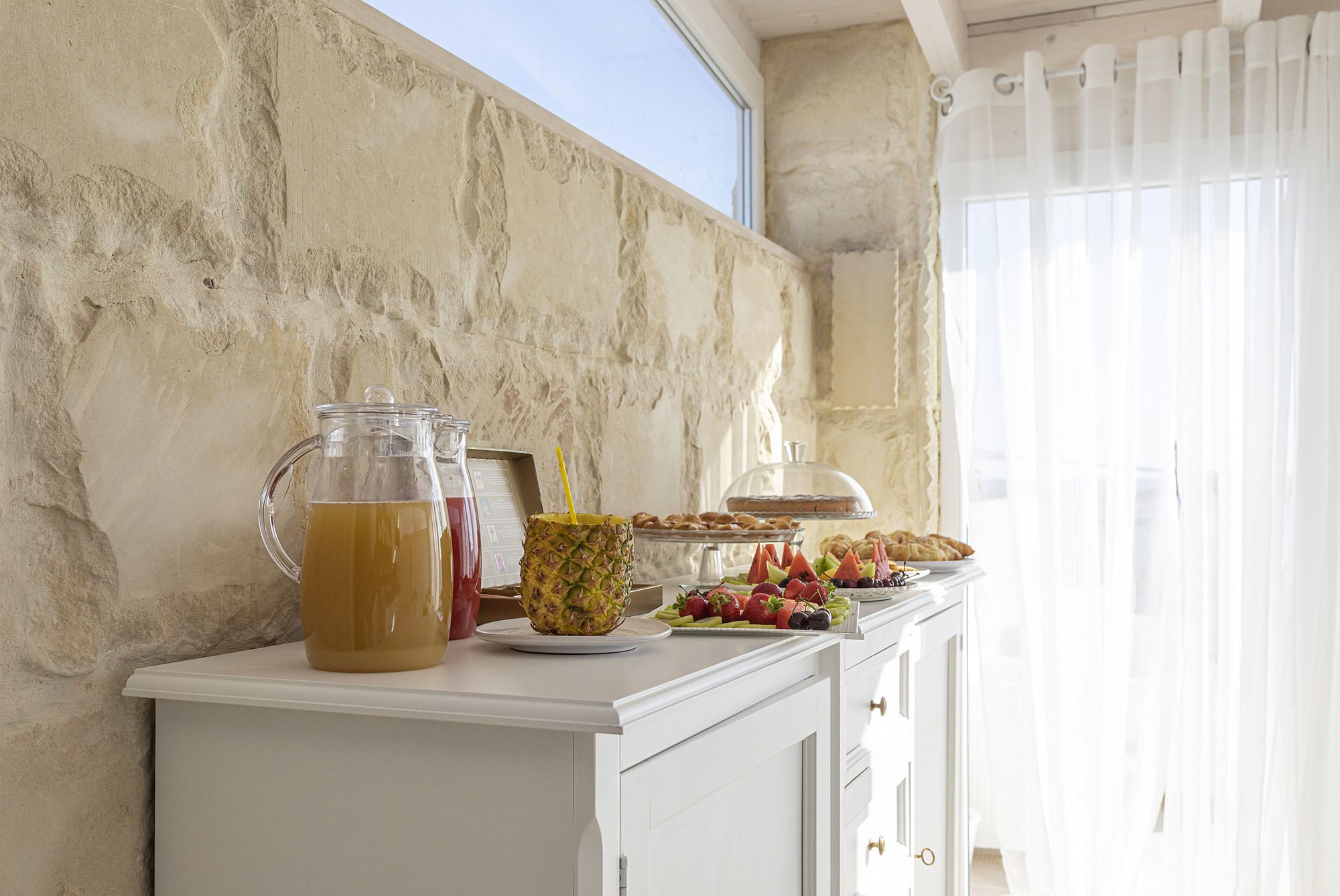 A breakfast buffet with a pitcher of orange juice, a pineapple, and various fruits and pastries on a white counter.