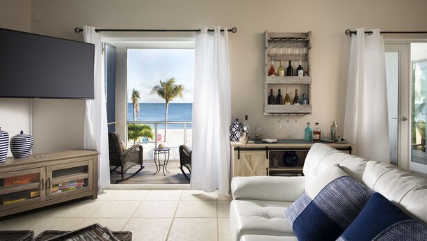 A living room with a white sofa, a flat-screen TV, a wooden cabinet, and a view of the beach.