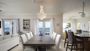 A dining area with a wooden table and white upholstered chairs, a chandelier, and a view of the ocean through large windows.