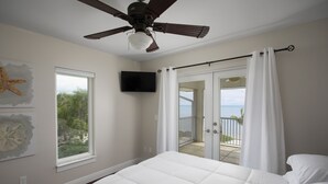 A bedroom with a bed, a ceiling fan, a TV, a window, and a balcony with a view of the ocean.