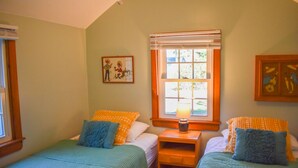 A bedroom with two beds, a window with blinds, a bedside table with a lamp, and a framed picture on the wall. - Charming 2-Bedroom Cottage Just 2/10 Mile from Sea Street Beach! (Dennis Port)