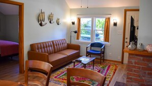 A living room with a brown sofa, a blue chair, a colorful rug, and a brick fireplace. - Charming 2-Bedroom Cottage Just 2/10 Mile from Sea Street Beach! (Dennis Port)