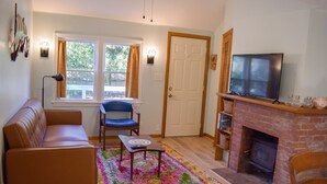 A living room with a sofa, a chair, a coffee table, a fireplace, a television, and a window with curtains. - Charming 2-Bedroom Cottage Just 2/10 Mile from Sea Street Beach! (Dennis Port)