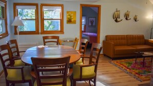 A dining area with a round wooden table and chairs, a sofa, and a colorful rug. - Charming 2-Bedroom Cottage Just 2/10 Mile from Sea Street Beach! (Dennis Port)