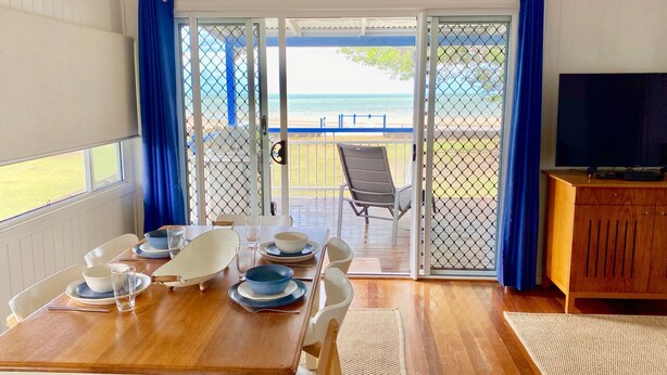 A dining area with a table set for four, a TV on a wooden cabinet, and a view of the beach through a glass door. - Burrum Beach House-Waterfront-Private Jetty-2BR (Burrum Heads)