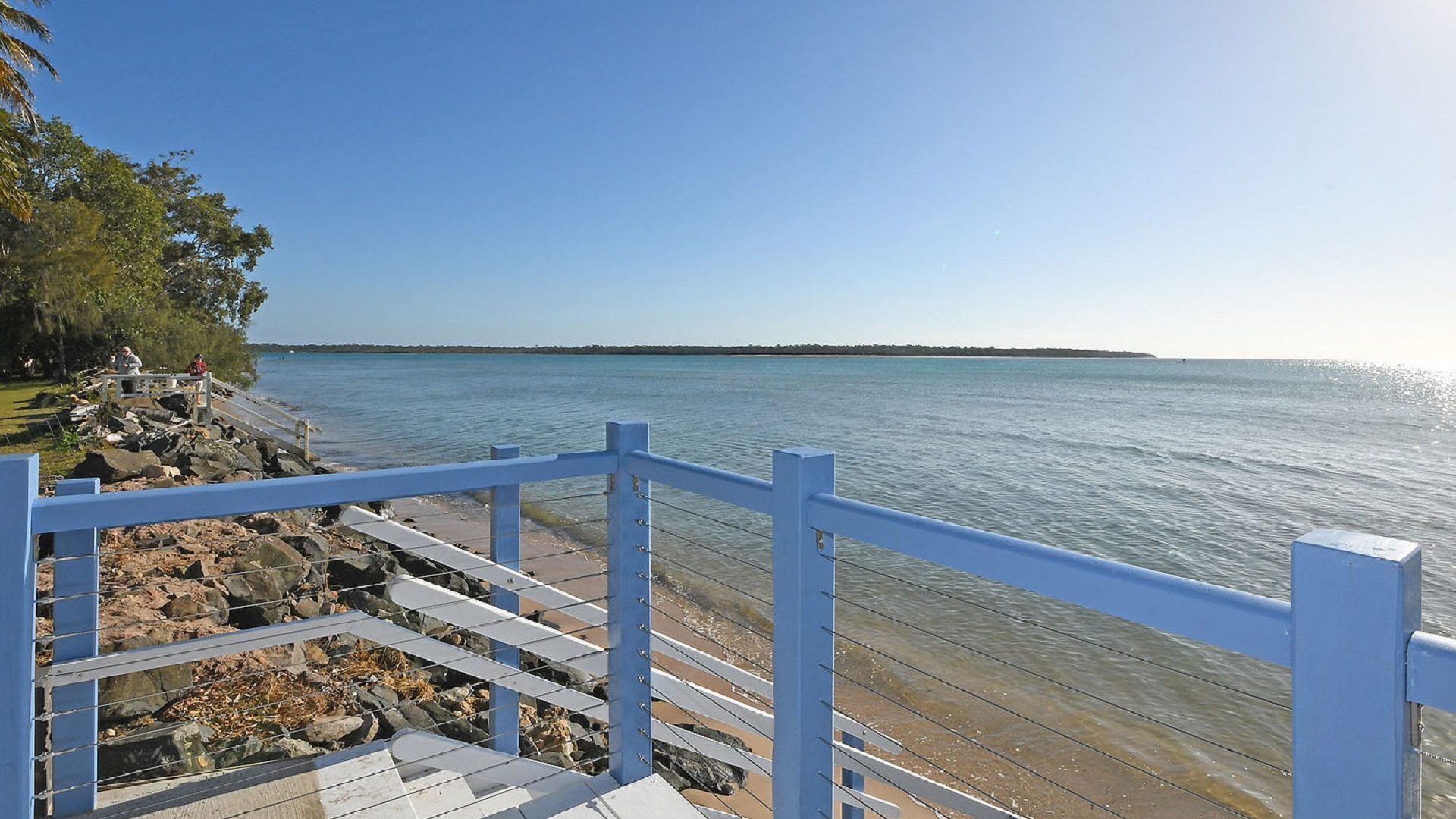 A wooden deck with a railing overlooking a calm body of water, with a rocky shoreline and trees in the background.