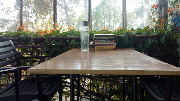An indoor seating area with a wooden table, chairs, and a stack of books. - Elation Guest House & Musafir Cafe (Ranikhet)
