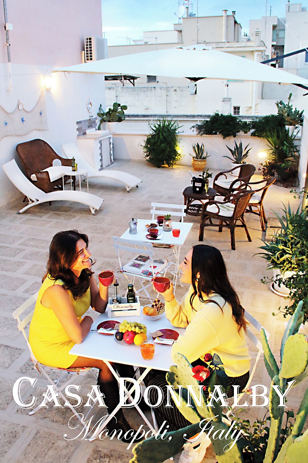 An outdoor patio with white furniture, a table set with fruits and drinks, and a sign reading "Casa Donnalby Monopoli, Italy".