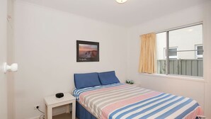 A bedroom with a bed, a bedside table, a nightstand, a window with curtains, and a framed picture on the wall. - Beachside Shoal Bay Unit 6 (Shoal Bay)