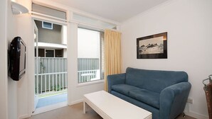 A room with a blue sofa, a white bench, a TV mounted on the wall, a window with curtains, and a framed picture. - Beachside Shoal Bay Unit 6 (Shoal Bay)
