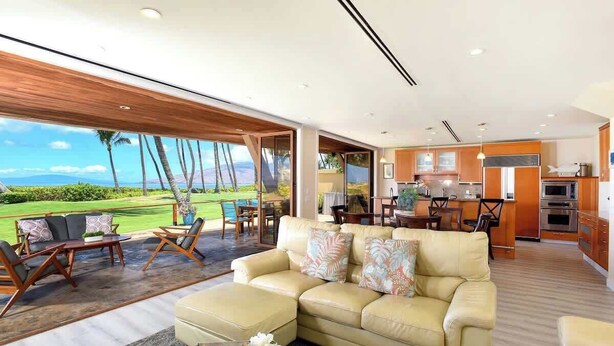 A spacious living area with a large window overlooking a golf course, a kitchen with modern appliances, and a cozy seating area with a sofa and chairs. - Aloha Villas # 2, Steps to Beach, Close to Golf & (Kihei)