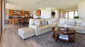 A spacious living area with a large beige sectional sofa, a glass coffee table, and a dining area with wooden furniture. - Aloha Villas # 2, Steps to Beach, Close to Golf & (Kihei)