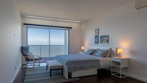 A modern bedroom with a large bed, a chair, a bench, a bedside table with a lamp, and a view through a large window. - Xenon urban apartments (Maputo)
