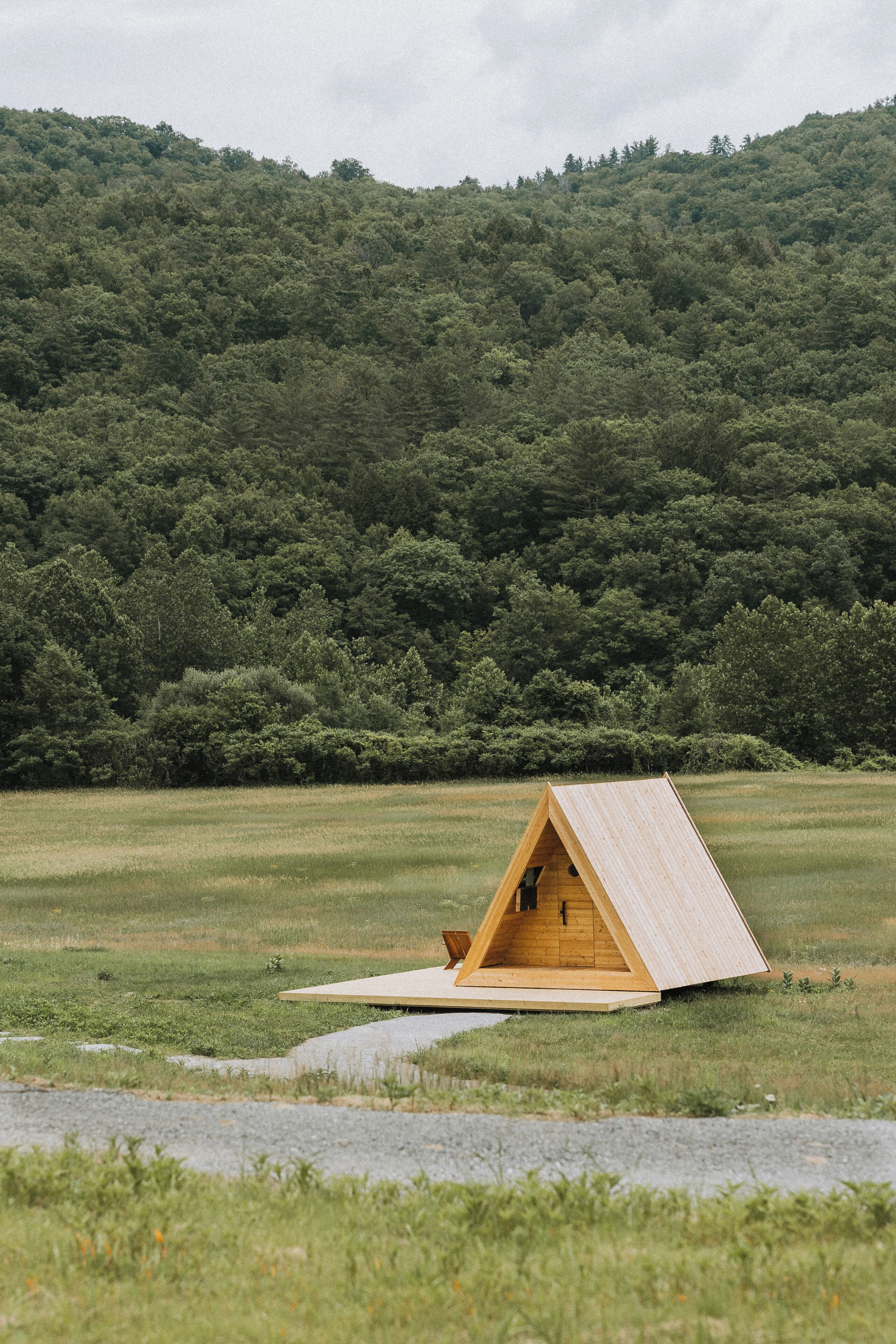 A wooden triangular structure in a grassy field with a forested hill in the background.
