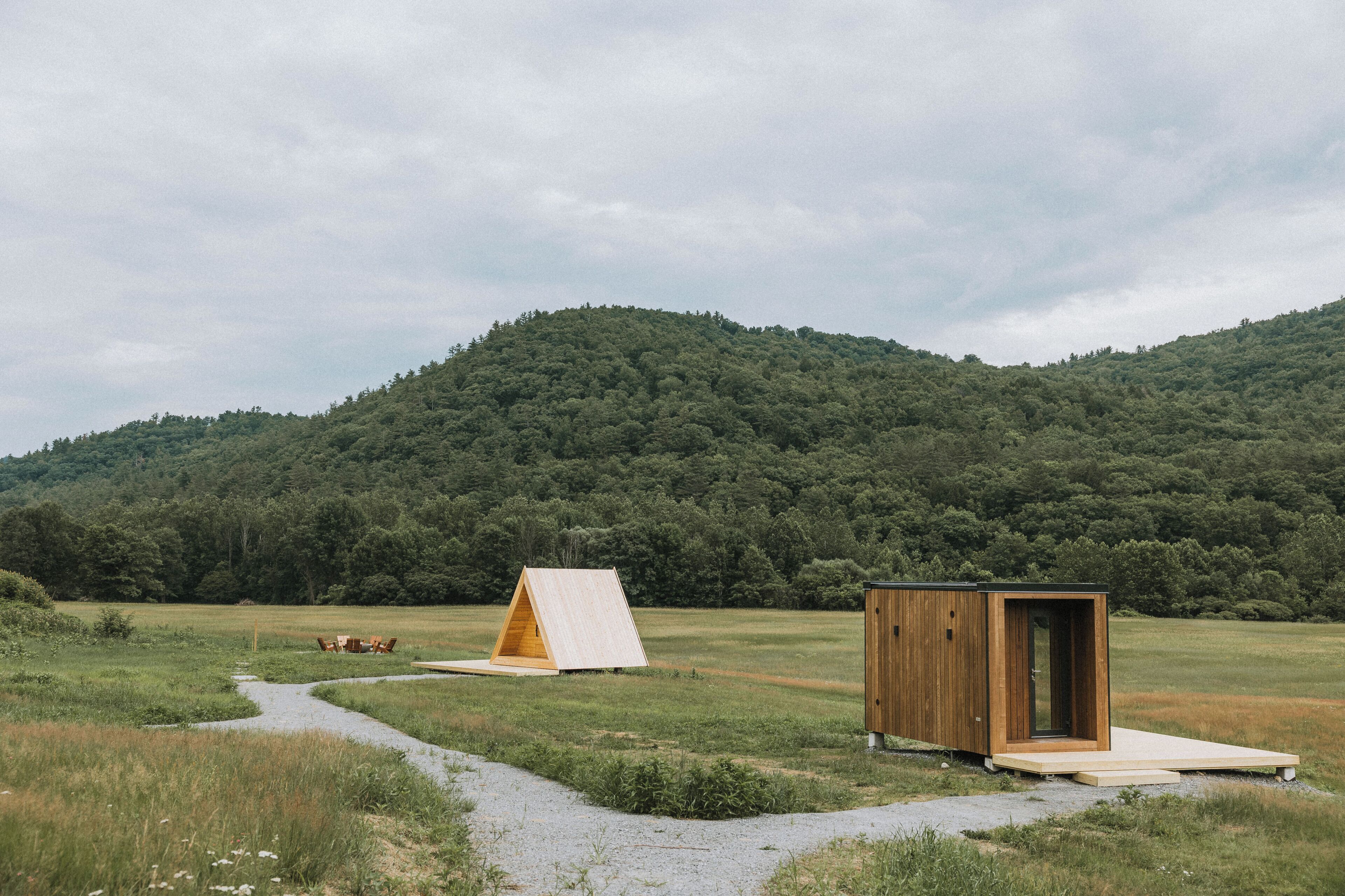 Een houten hutje met een klein terras, een tent en een grindpad dat leidt naar een grasveld met een berg op de achtergrond.