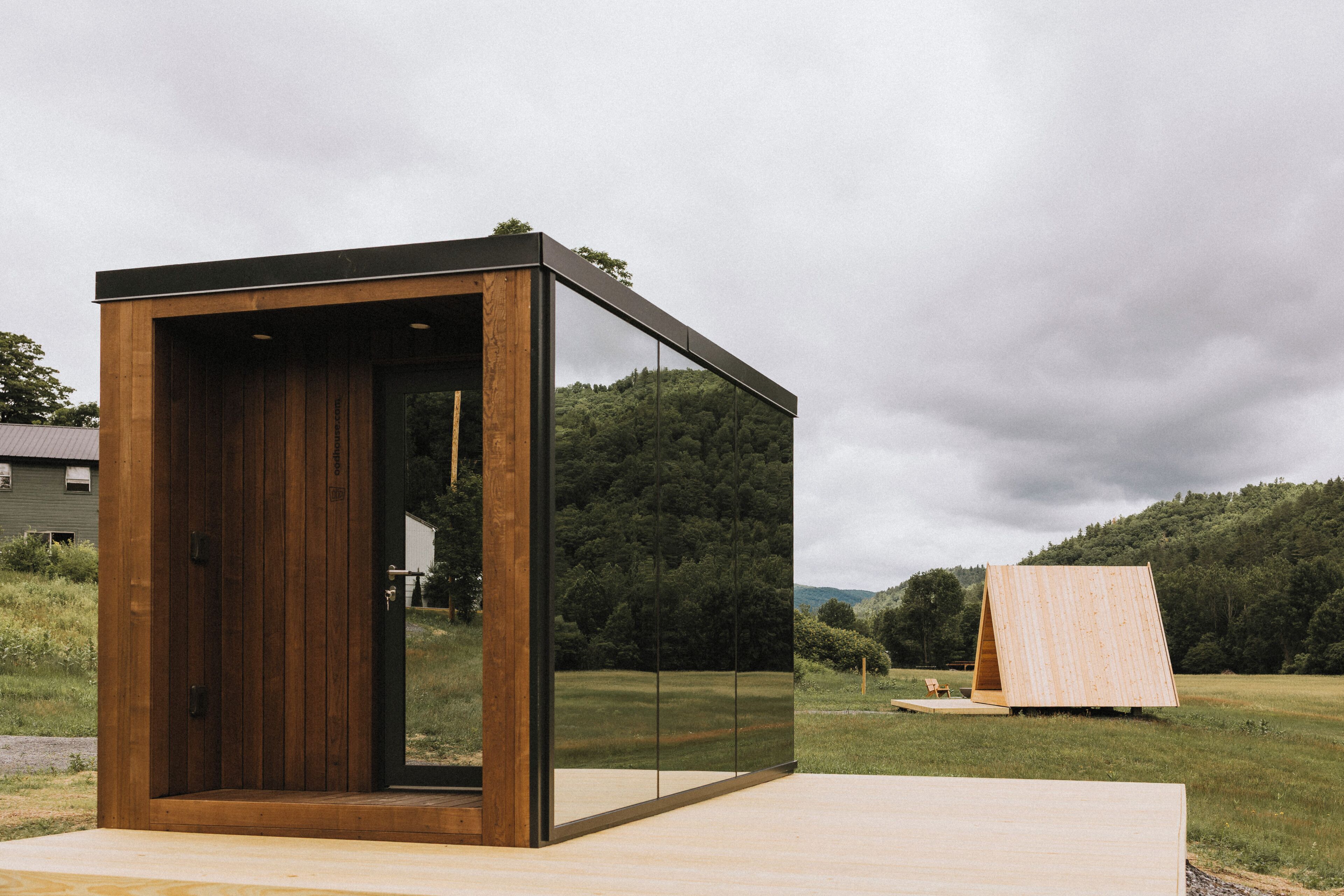 Ein modernes, minimalistisches Gebäude mit Holzfassade und Glaswänden, eingebettet in eine grüne Wiese mit einem bewaldeten Hügel im Hintergrund.