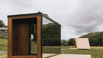Ein modernes, minimalistisches Gebäude mit Holzfassade und Glaswänden, eingebettet in eine grüne Wiese mit einem bewaldeten Hügel im Hintergrund.