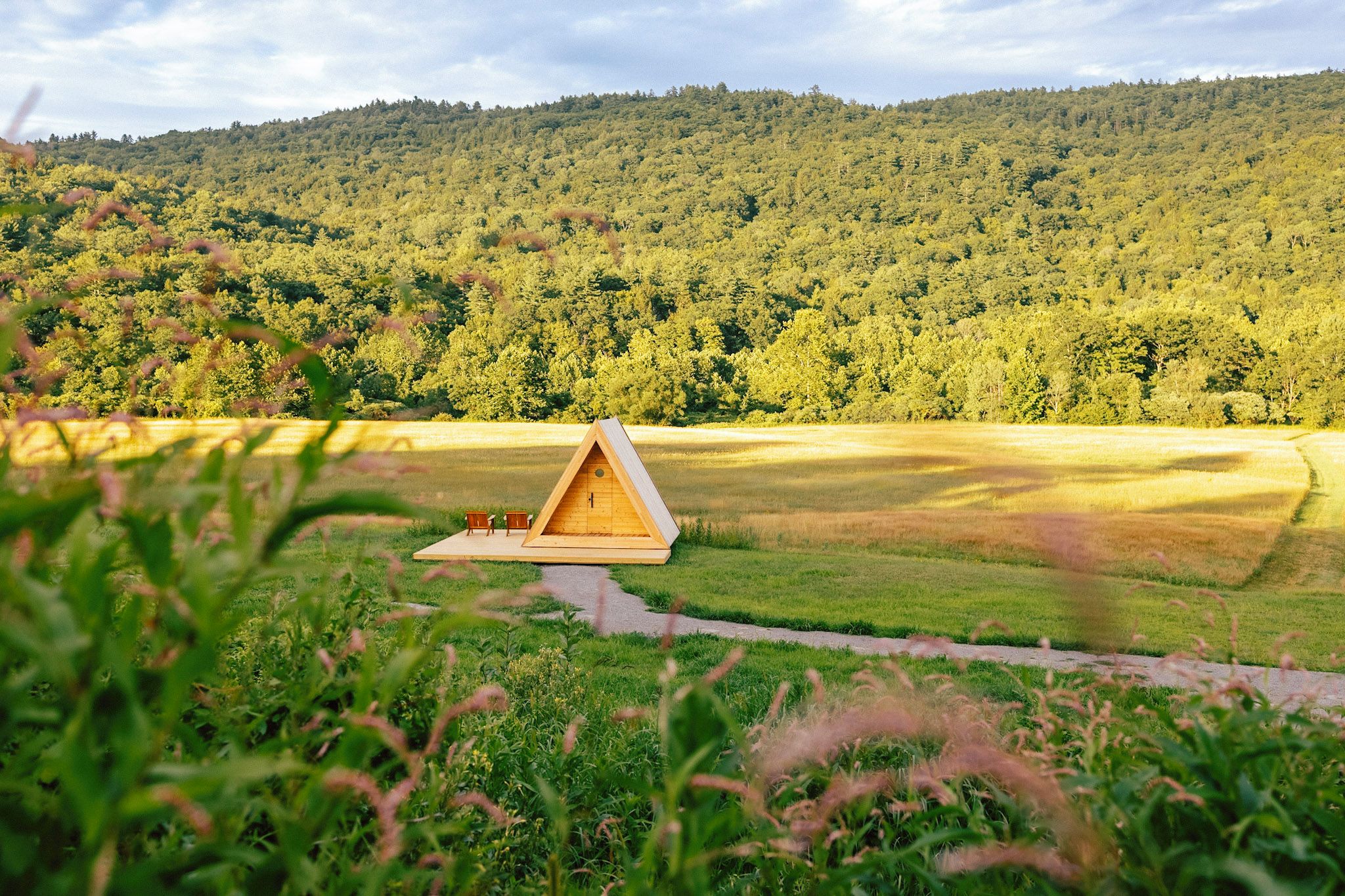 A wooden cabin with a triangular roof, situated in a grassy field with a forested hill in the background.