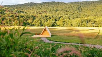 A wooden cabin with a triangular roof, situated in a grassy field with a forested hill in the background.