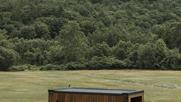 Een houten blokhut met één deur en een klein terras, gelegen in een grasveld met een bebost heuvel op de achtergrond.