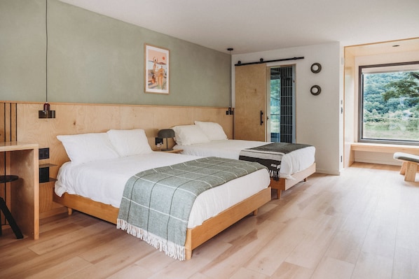 A modern bedroom with two beds, wooden headboards, a bench, a small table, and a large window with a view of greenery.