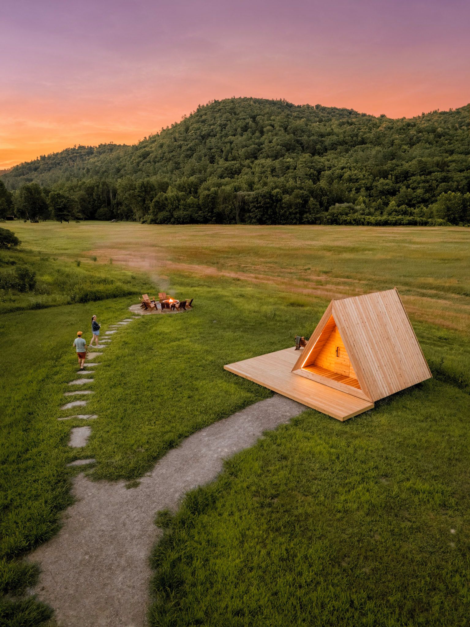 Una cabaña de madera con techo inclinado en un campo cubierto de hierba, un brasero con sillas y un sendero que lo conduce.