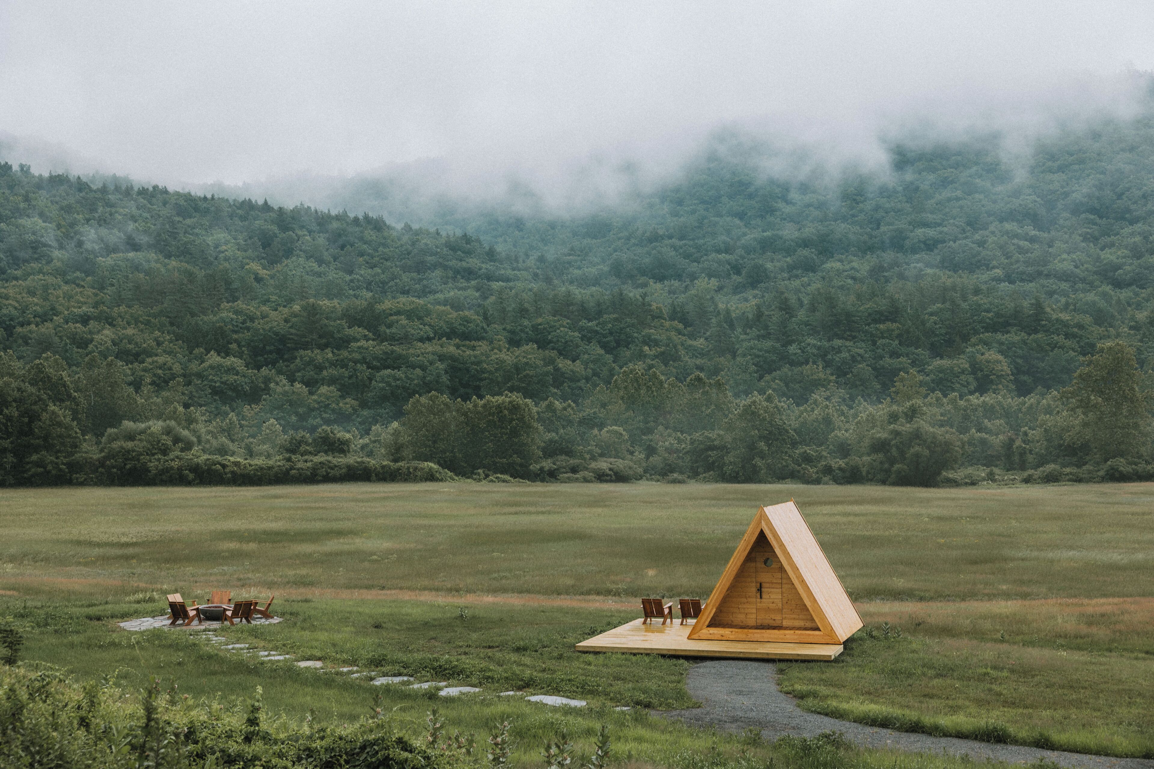 A wooden triangular cabin with a small deck and two chairs, surrounded by a grassy field and a forested hill.