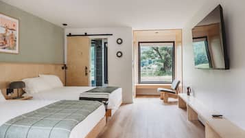 A modern hotel room with two beds, a desk, a chair, a TV, and a large window with a view of trees.