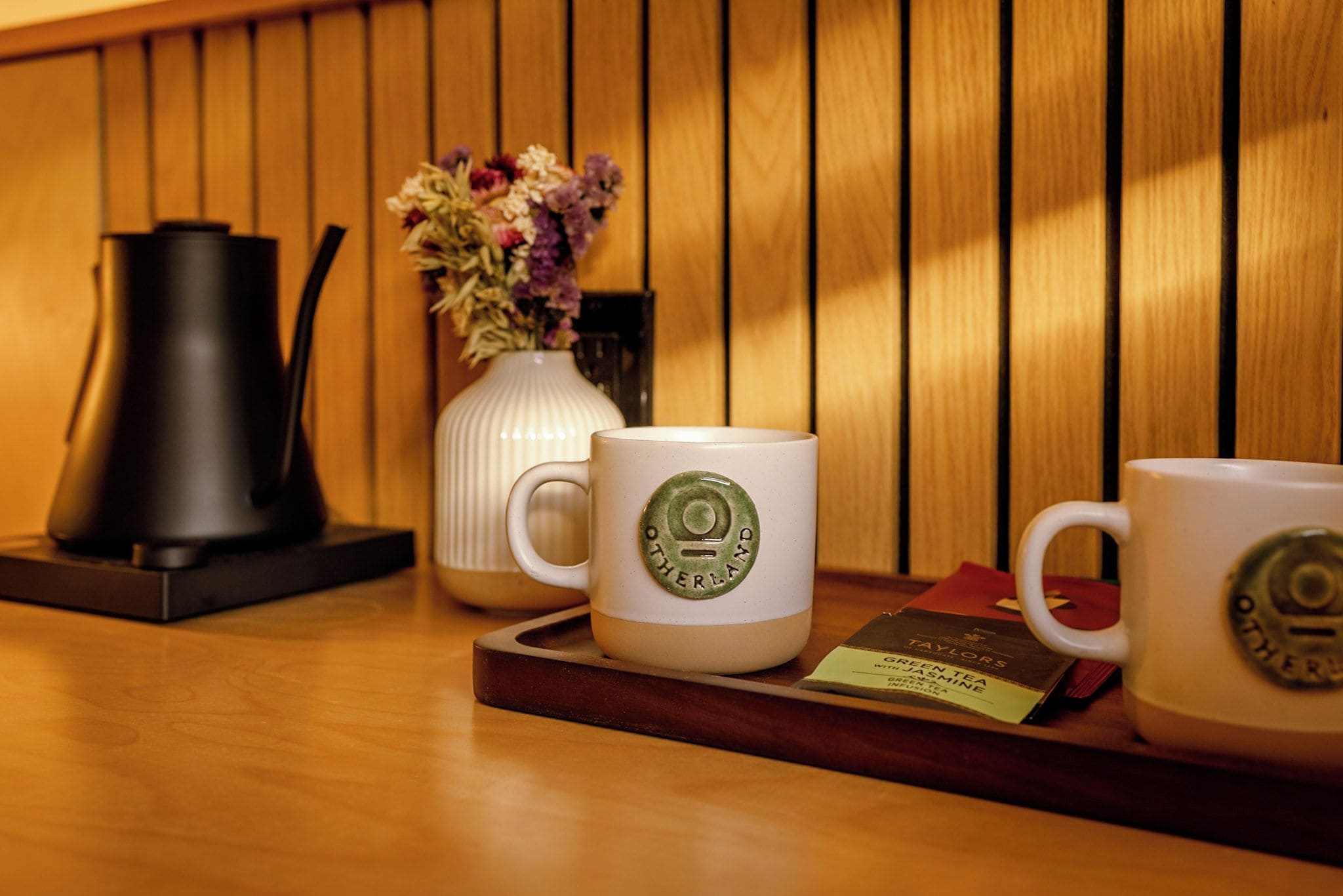A tray with two white mugs, a white vase with flowers, a black electric kettle, and a tray with a green tea package.