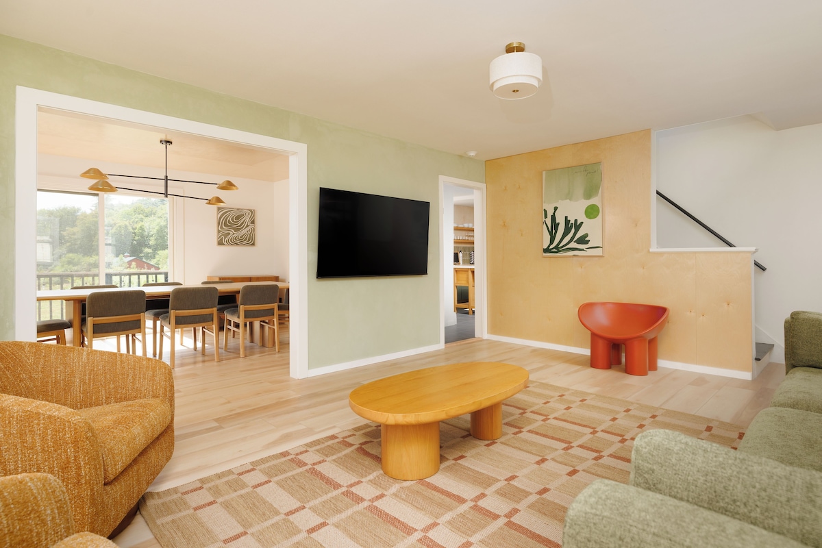 Un salon moderne avec un banc en bois, un coin repas avec des chaises, un téléviseur à écran plat et un escalier.