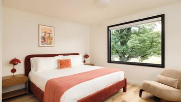 A bedroom with a large window, a bed with white linens, a red throw blanket, a side table with a lamp, and a framed artwork on the wall.