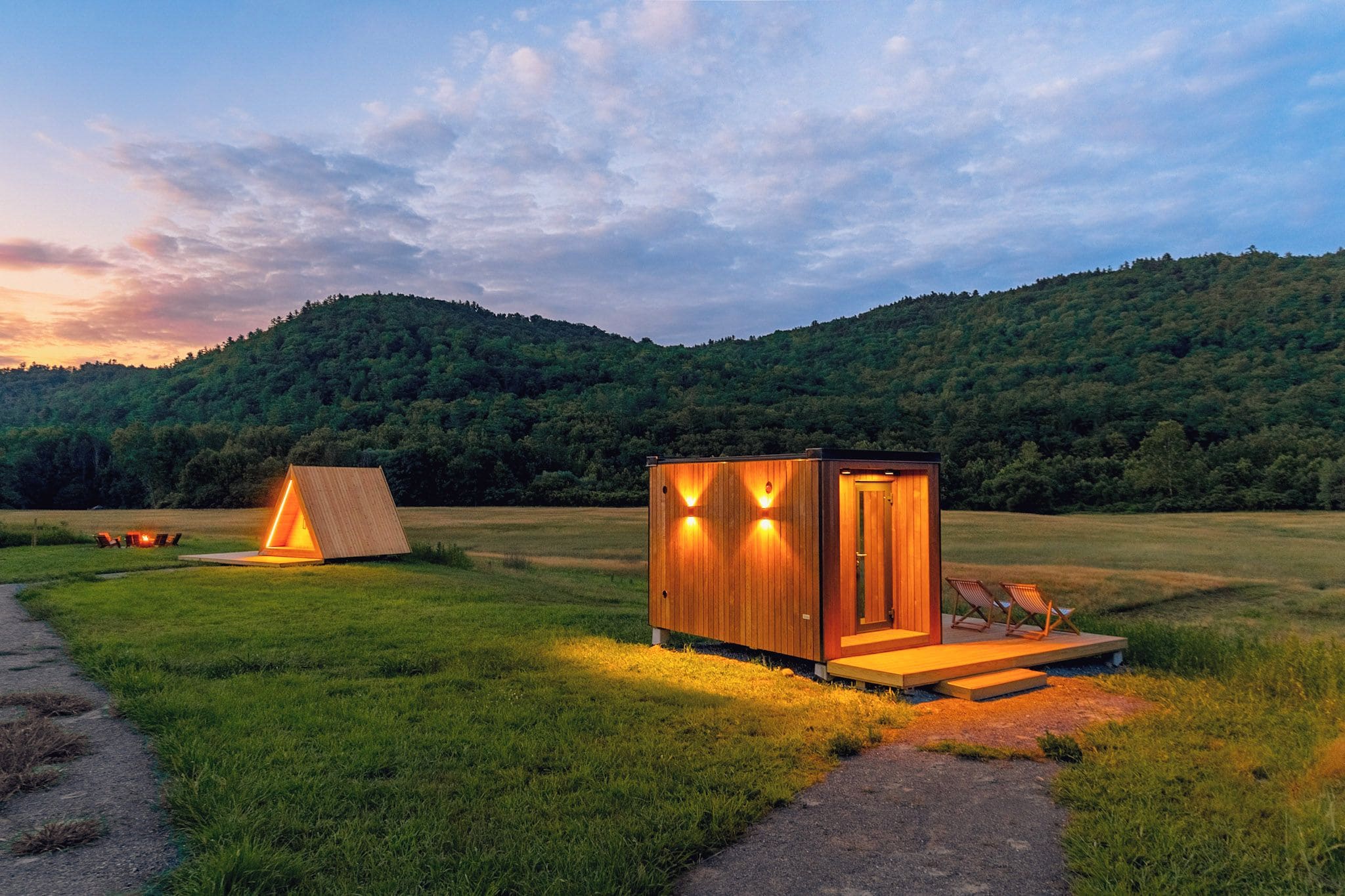 A campsite with a wooden cabin and a tent, surrounded by a grassy field and a forested hill.