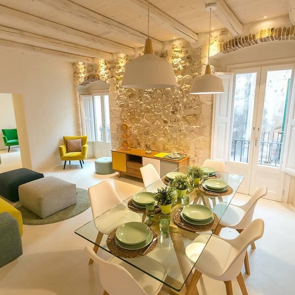 A dining area with a glass table set for four, surrounded by white chairs. A stone wall with a textured pattern is behind the table.