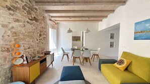 A modern living room with a stone wall, wooden ceiling beams, a dining area with white chairs, and a yellow sofa with a patterned pillow. - Ortigia Luxury Apartment w/ Sea View & Terraces (Syracuse)