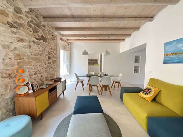 Una sala moderna con pared de piedra, vigas de madera en el techo, una zona de comedor con sillas blancas y un sofá amarillo con un cojín estampado.