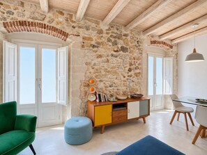 Ein Zimmer mit einer Steinwand, Holzbalken, einem grünen Sessel, einem blauen Hocker, einem gelb-weißen Schrank und einem Glastisch mit Stühlen.