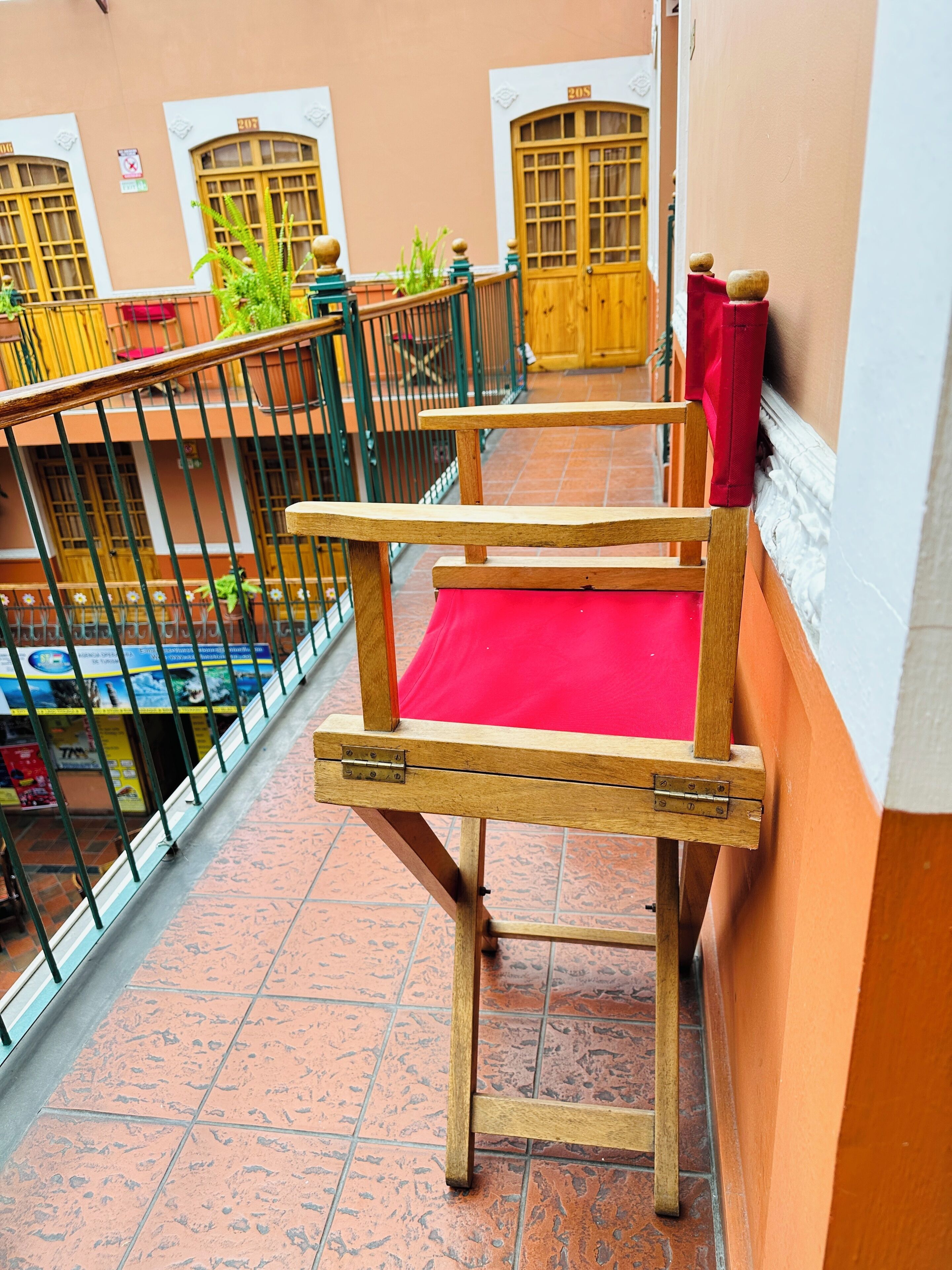 Una silla de madera con un cojín rojo sobre un suelo de baldosas, una barandilla de metal y la fachada de un edificio con ventanas numeradas.