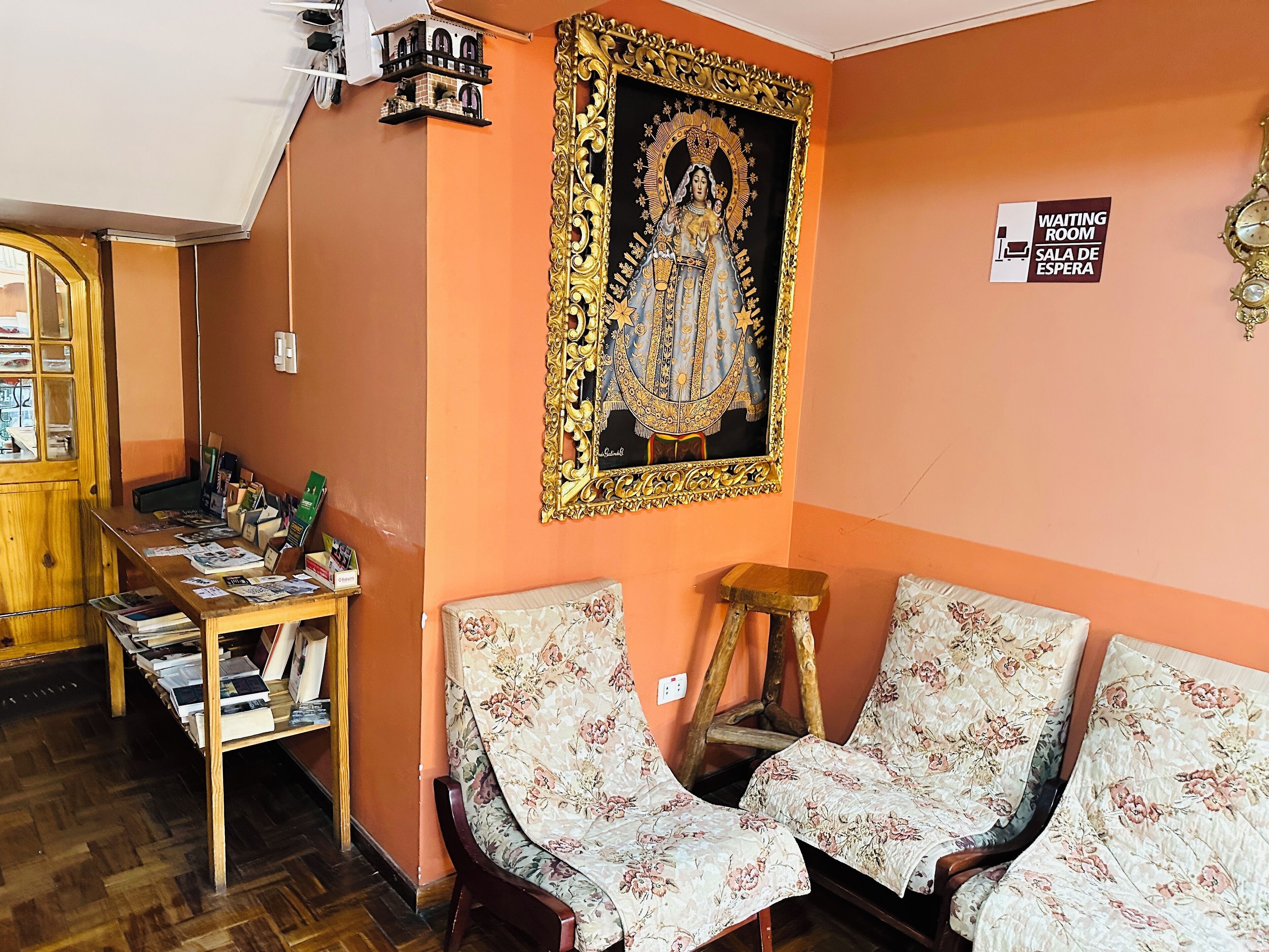 Ein Wartezimmer mit einem religiösen Gemälde, einem Holztisch mit Zeitschriften und zwei stuhlartigen Sitzgelegenheiten mit Blumenmuster.