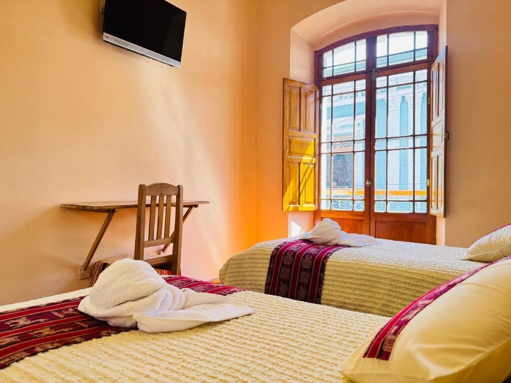 A hotel room with two beds, a TV mounted on the wall, a window with a view of buildings, and a table with a chair.
