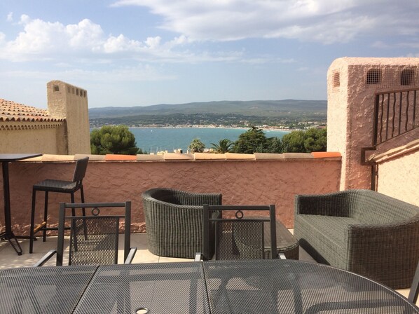 A terrace with wicker furniture, a table, and a view of the sea and distant landscape.