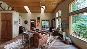 Un salon spacieux avec un plafond haut, des poutres en bois et de grandes fenêtres offrant une vue sur la forêt.