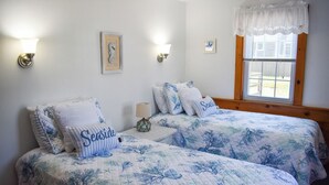 Une chambre à coucher avec deux lits, chacun recouvert d’une courtepointe et de coussins à motifs floraux bleus et blancs, et portant le mot « Seaside » imprimé dessus. On y trouve des lampes fixées au mur et une fenêtre agrémentée d’un lambrequin blanc.