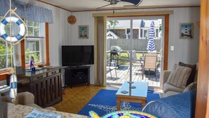 Un salon équipé d’une télévision et d’un ventilateur de plafond, un coin repas avec des sièges extérieurs, et une vue sur le jardin arrière.